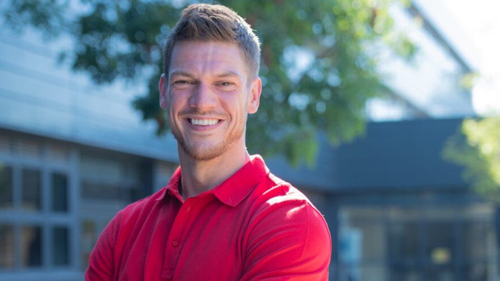 Jason Grant is Scotland's new Period Dignity Officer. He's wearing a red polo in front of a tree and building