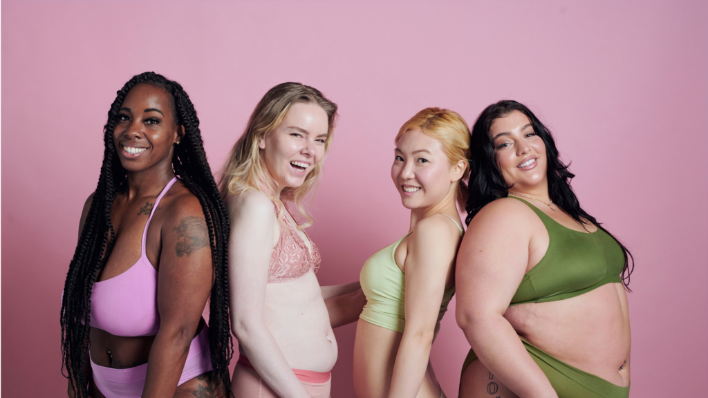Four women in lingerie of different colors and sizes stand before a pink wall. Blog: how to connect with your body