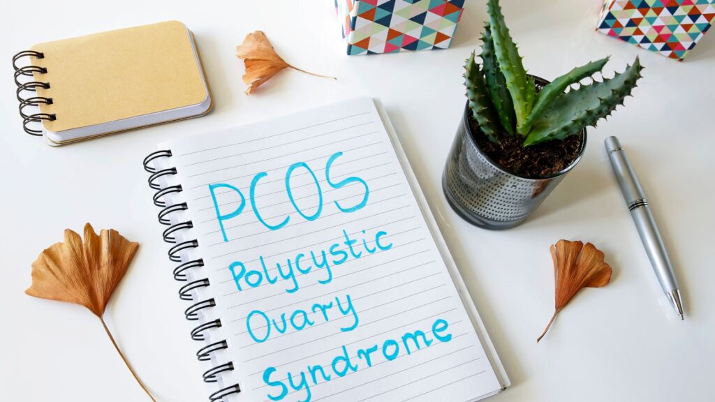 Notebook on a desktop surrounded by a succulent, flower, pen with the words: PCOS Polycystic Ovary Syndrome written in blue