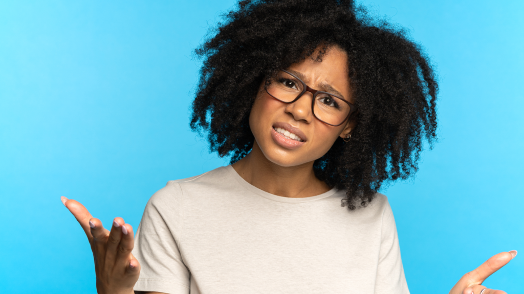 Black woman with curly hair wearing an oatmeal colored shirt and glasses with her hands up in frustration