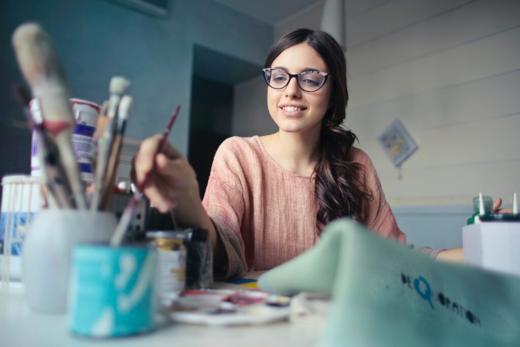 Woman wearing glasses, dipping a paint brush into water while working on a painting. Blog post about embracing the journey of creativity.