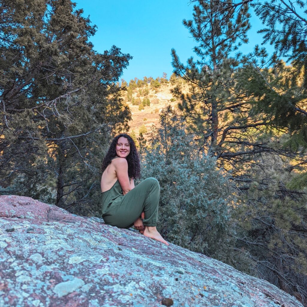 Image of Colleen Howell sitting on a big rock with trees in the background. She has long dark curly hair and is wearing a green romper. This is a blog post Colleen wrote about how the menstrual cycle phases correlate with feminine archetypes. 