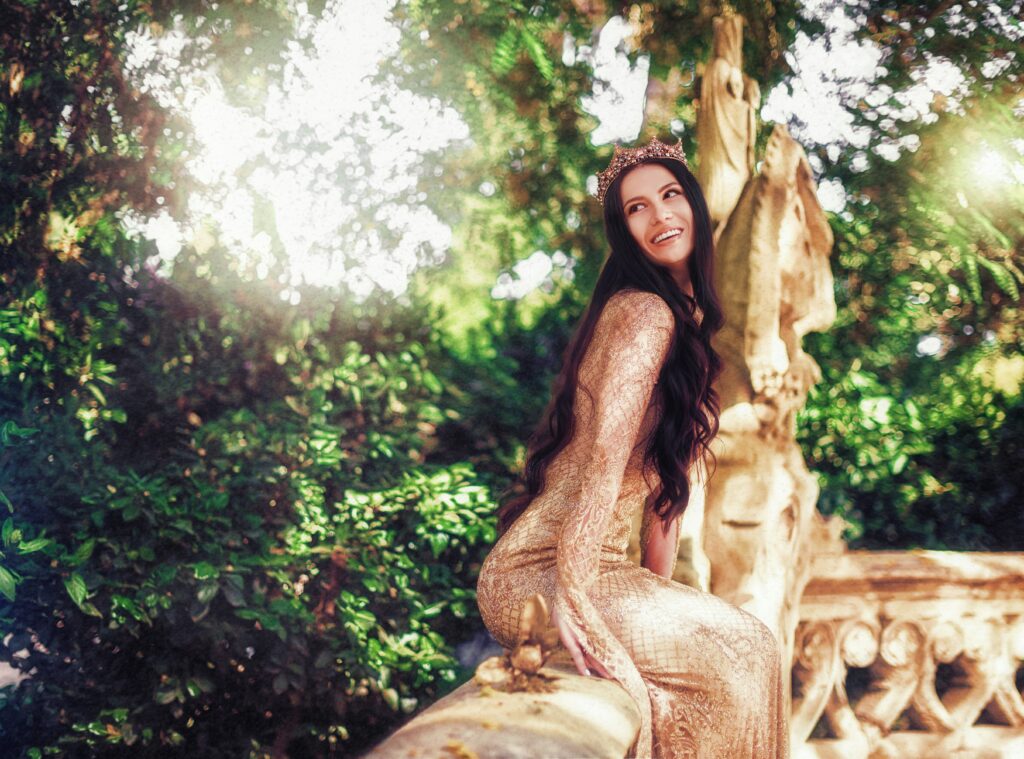 Woman with long dark hair dressed like a queen in long golden dress and a crown. She's sitting on a balcony ledge with greenery behind her. The queen is a common feminine archetype that is often associated with the ovulatory phase.